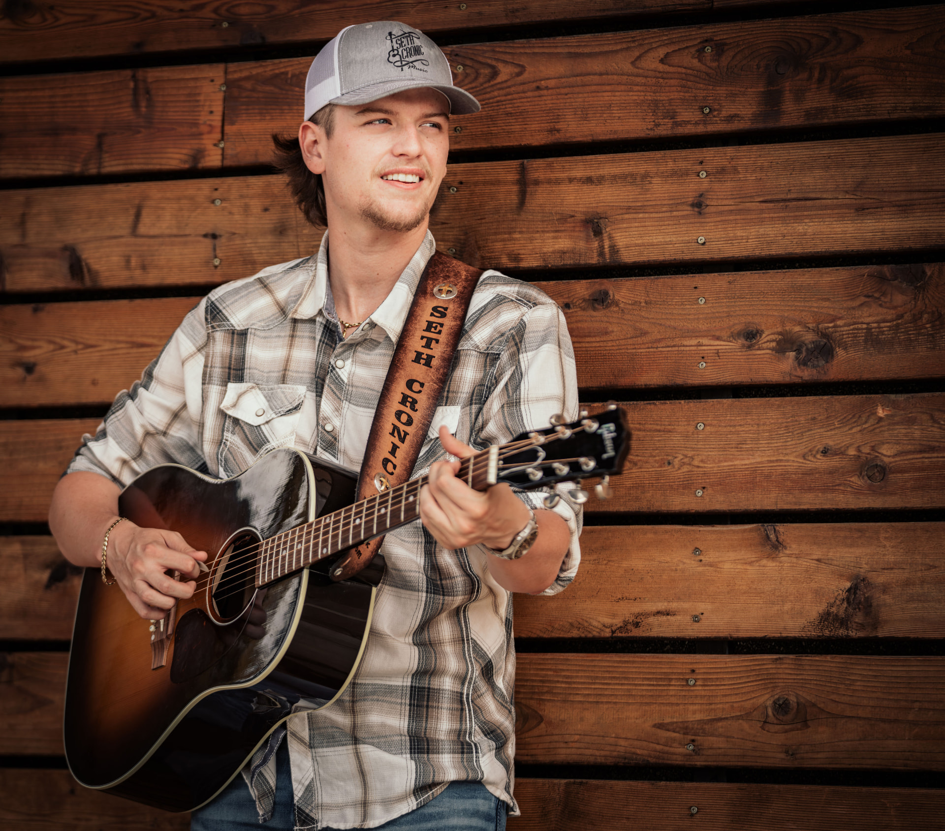 Seth Cronic smiling with his acoustic guitar against a wooden wall, wearing a Seth Cronic Music cap
