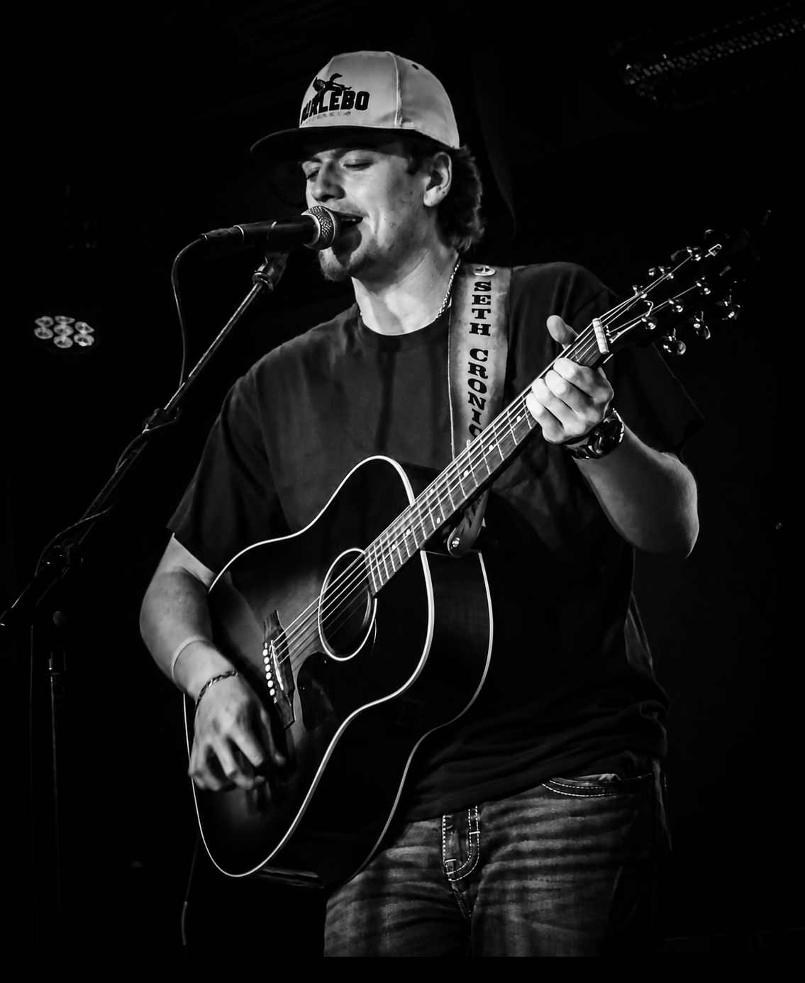 Seth Cronic singing and playing guitar on stage wearing a Seth Cronic Music cap